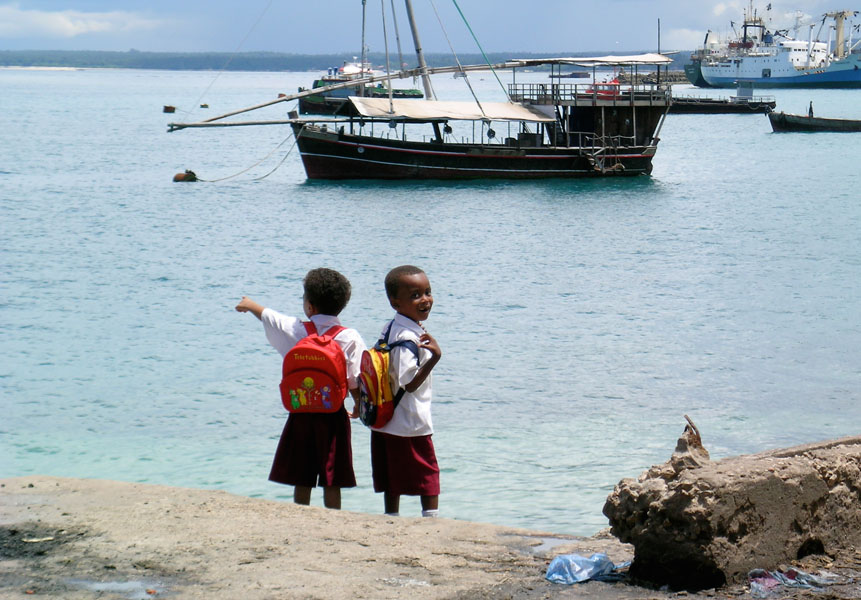 Two school kids with uniform and backpacks are standing in the harbor of Zanzibar City and point to the ships