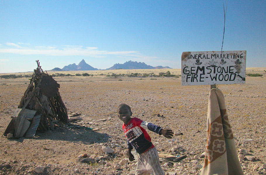 A child in Namibia sells gem stones and firewood on the street under a sign labeled "Mineral Marketing". In the background one can see the "Spitzkoppe" mountains