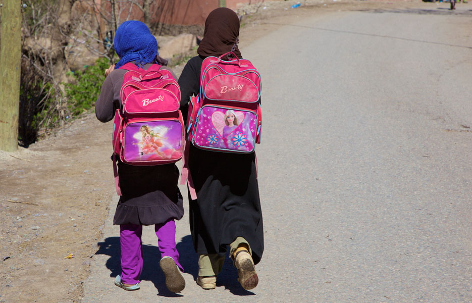 Two schoolgirls wearing headscarves and beauty schoolbags on their backs walk along a road in Imlil / Marrakech / Morocco